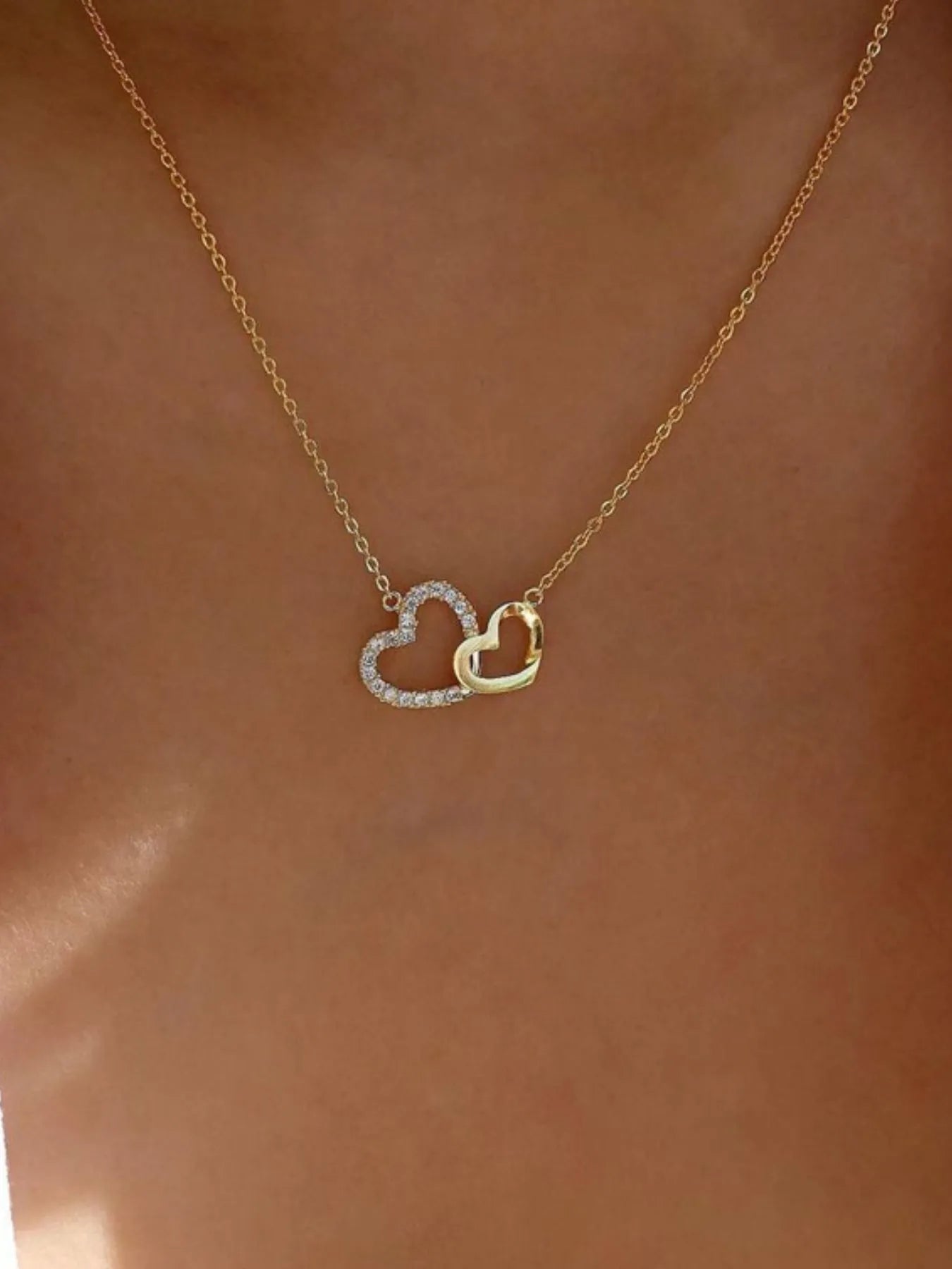 Double Heart Pendant Necklace with gold chain and intertwined hearts, one encrusted with zircon stones, worn on a model's neck.