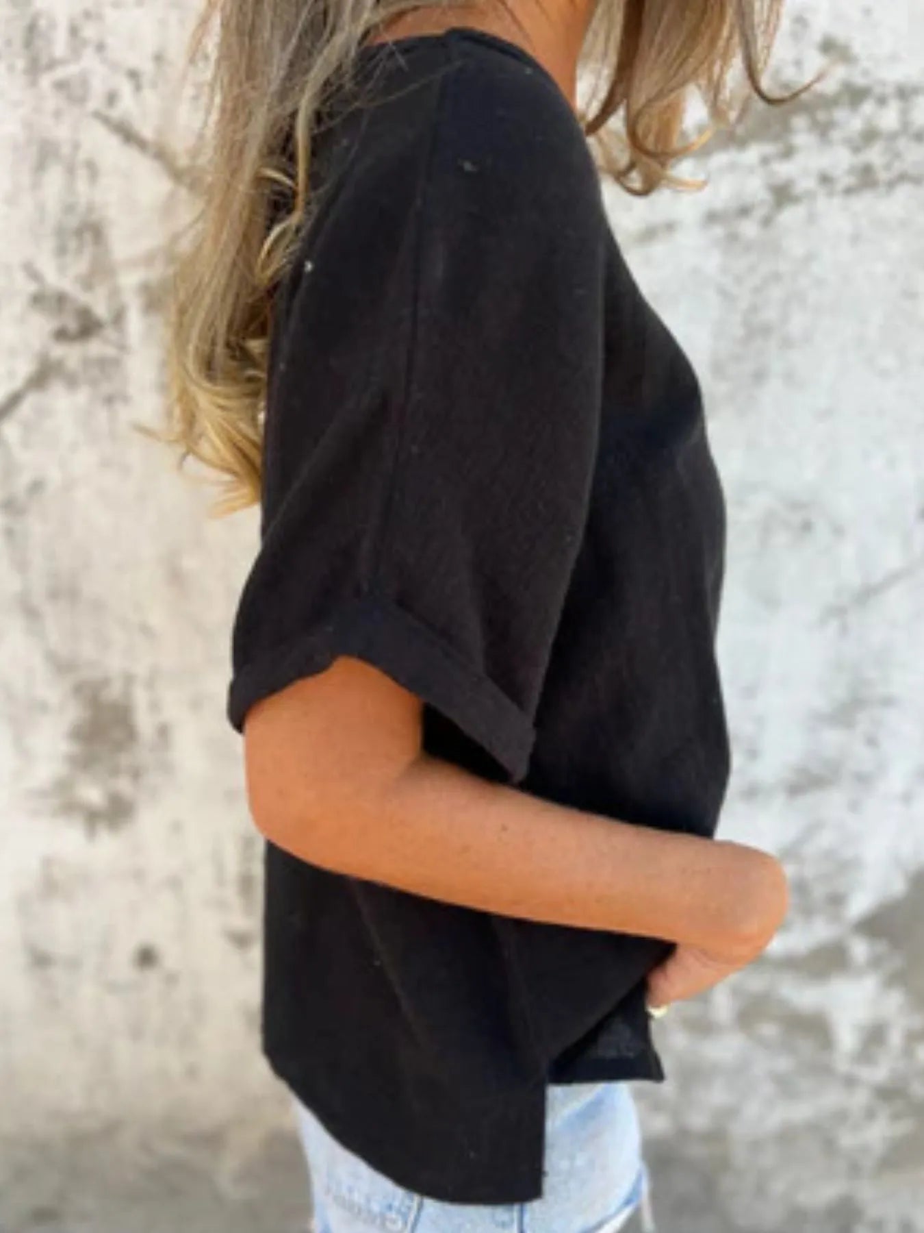 Side profile of black Women's Oversized Essential Tee with rolled sleeves and curved hem, styled with casual shorts.