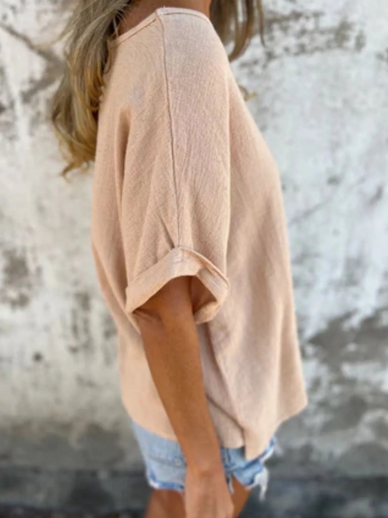 Side view of beige Women's Oversized Essential Tee with rolled sleeves and a loose silhouette, styled with denim shorts.