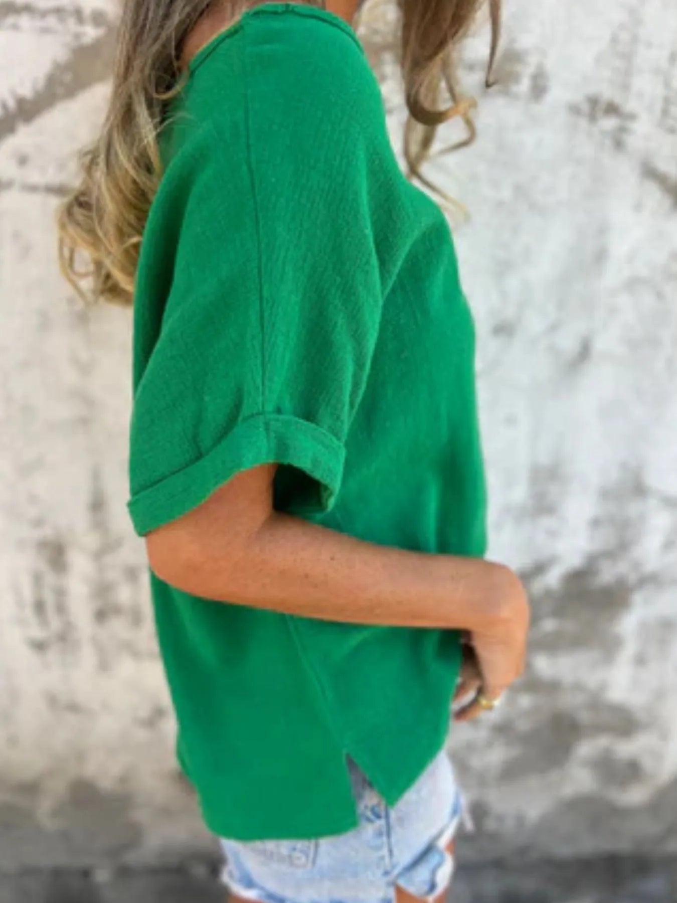 Side view of green Women's Oversized Essential Tee featuring rolled sleeves and side slit hem, styled with denim shorts.