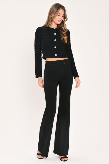 A woman modeling a black ribbed long-sleeve cropped cardigan with silver buttons and matching black high-waisted flared pants, standing against a plain off-white studio background.