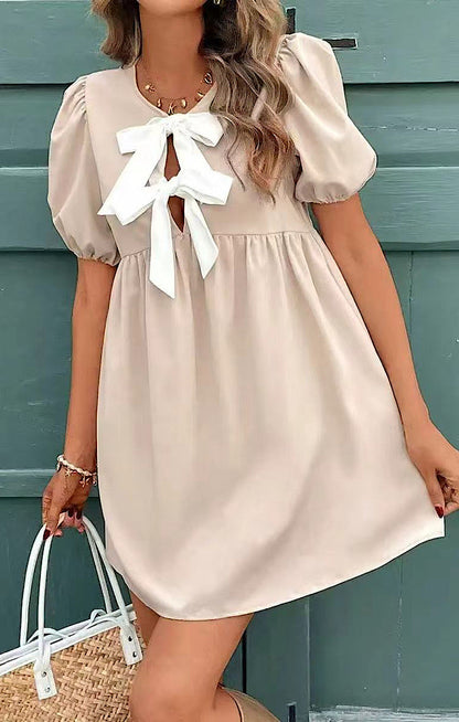 Woman wearing a beige dress with a white bow in front of a green wooden wall.