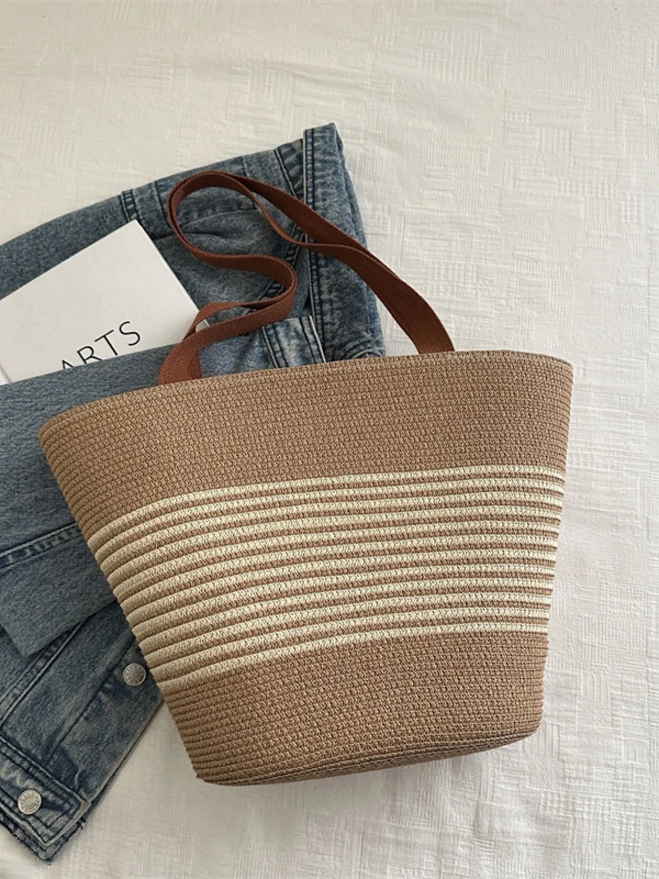 Beige and cream striped woven tote bag with brown handles, laid flat on white surface with denim jacket. Stylish and sustainable.