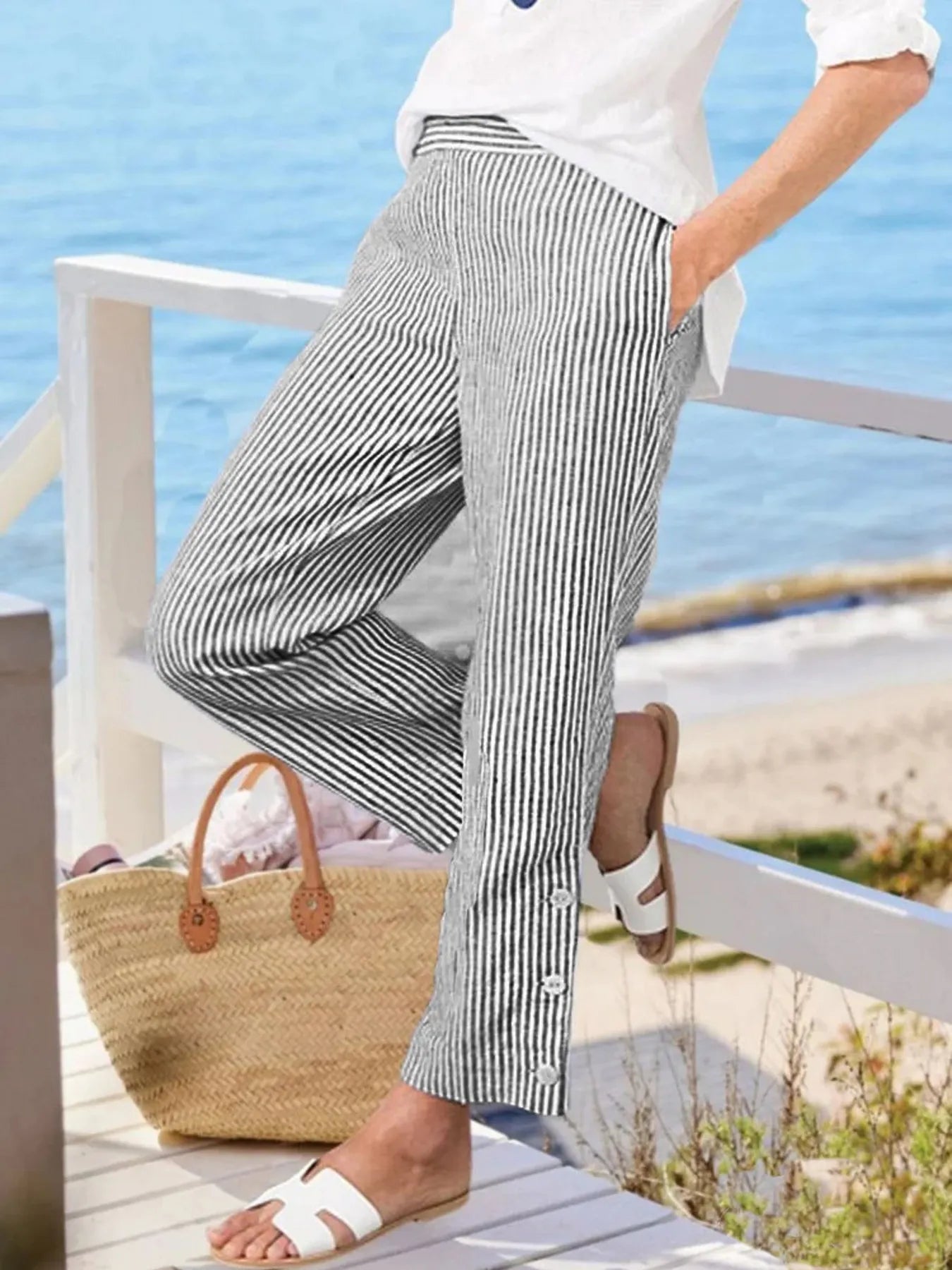 Madison Striped Button-Hem Pants in black and white, styled by the beach with a straw tote. Features button accents at the hem.