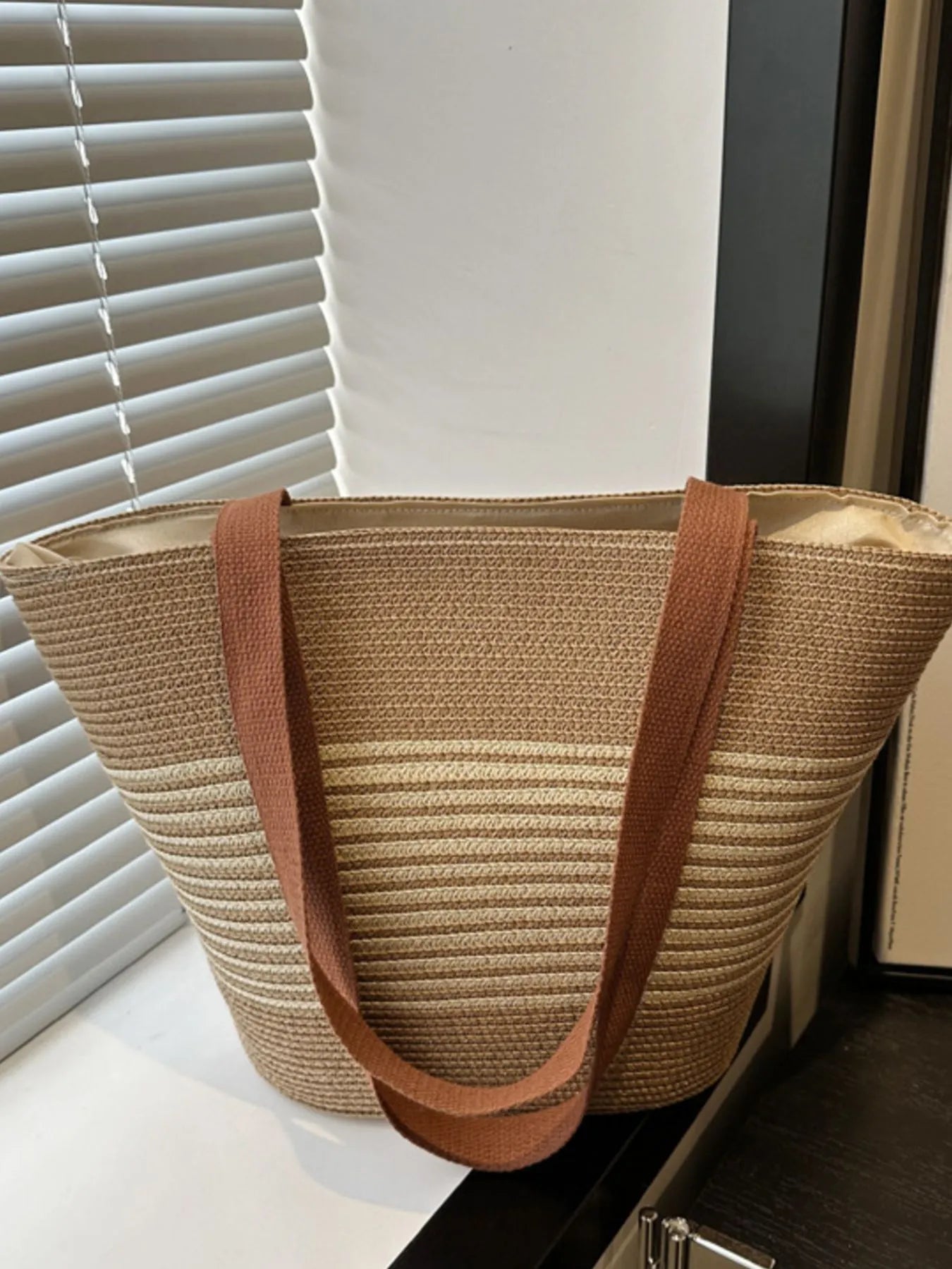 Back view of striped woven medium size tote bag in beige and cream tones with brown straps, placed on dark surface near window.