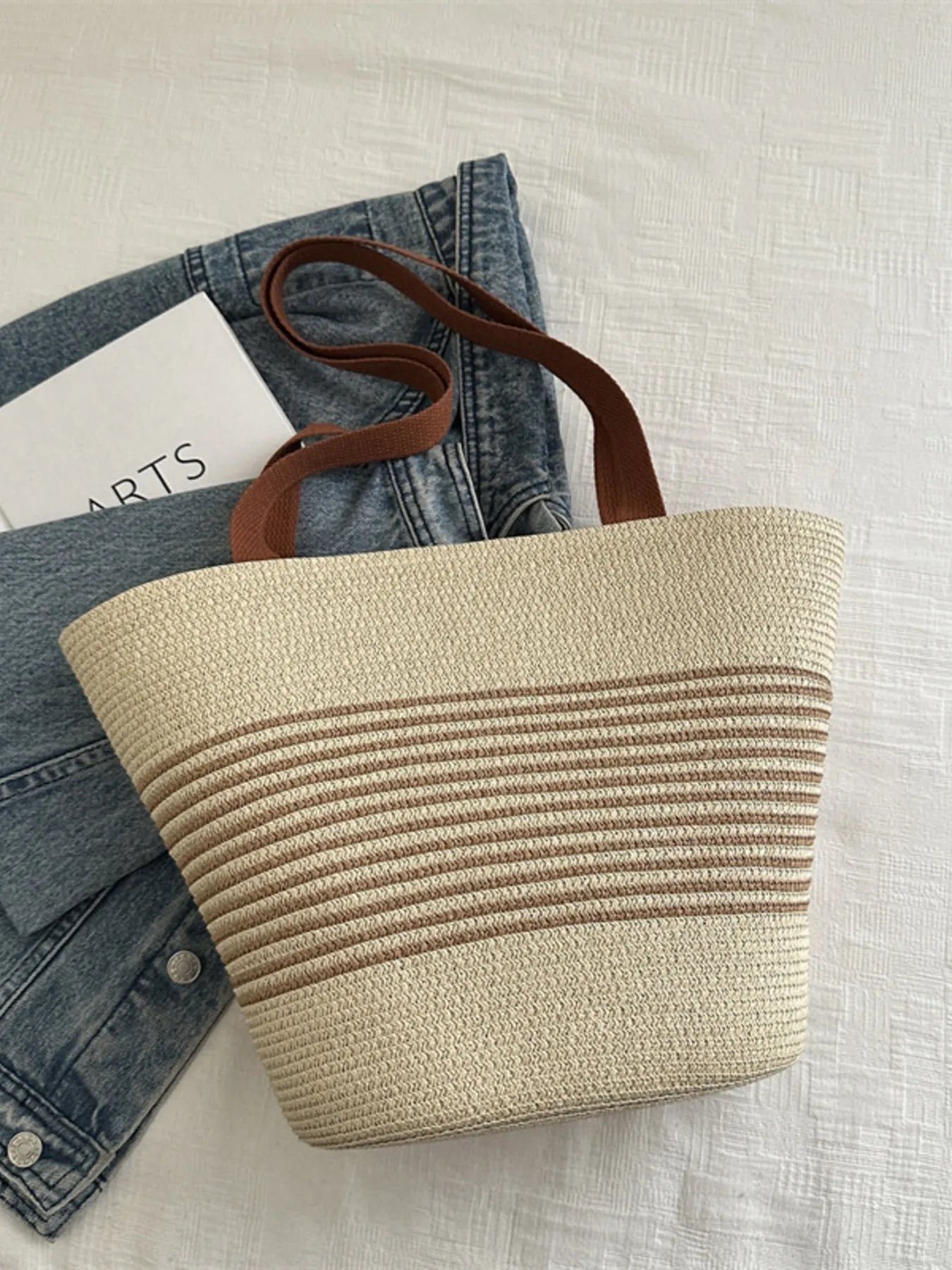 Light cream and tan striped woven tote bag with brown handles, placed on white fabric with denim jacket. Chic and eco-conscious design.