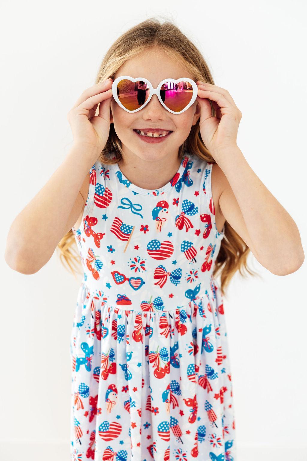 Young girl wearing a dress with American flag pattern and heart-shaped sunglasses.